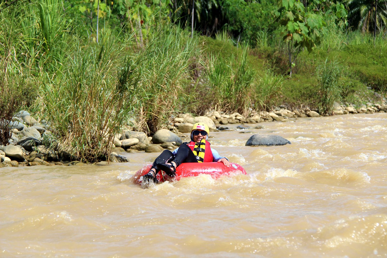 A guest floats past tall grass and river rocks in her tube